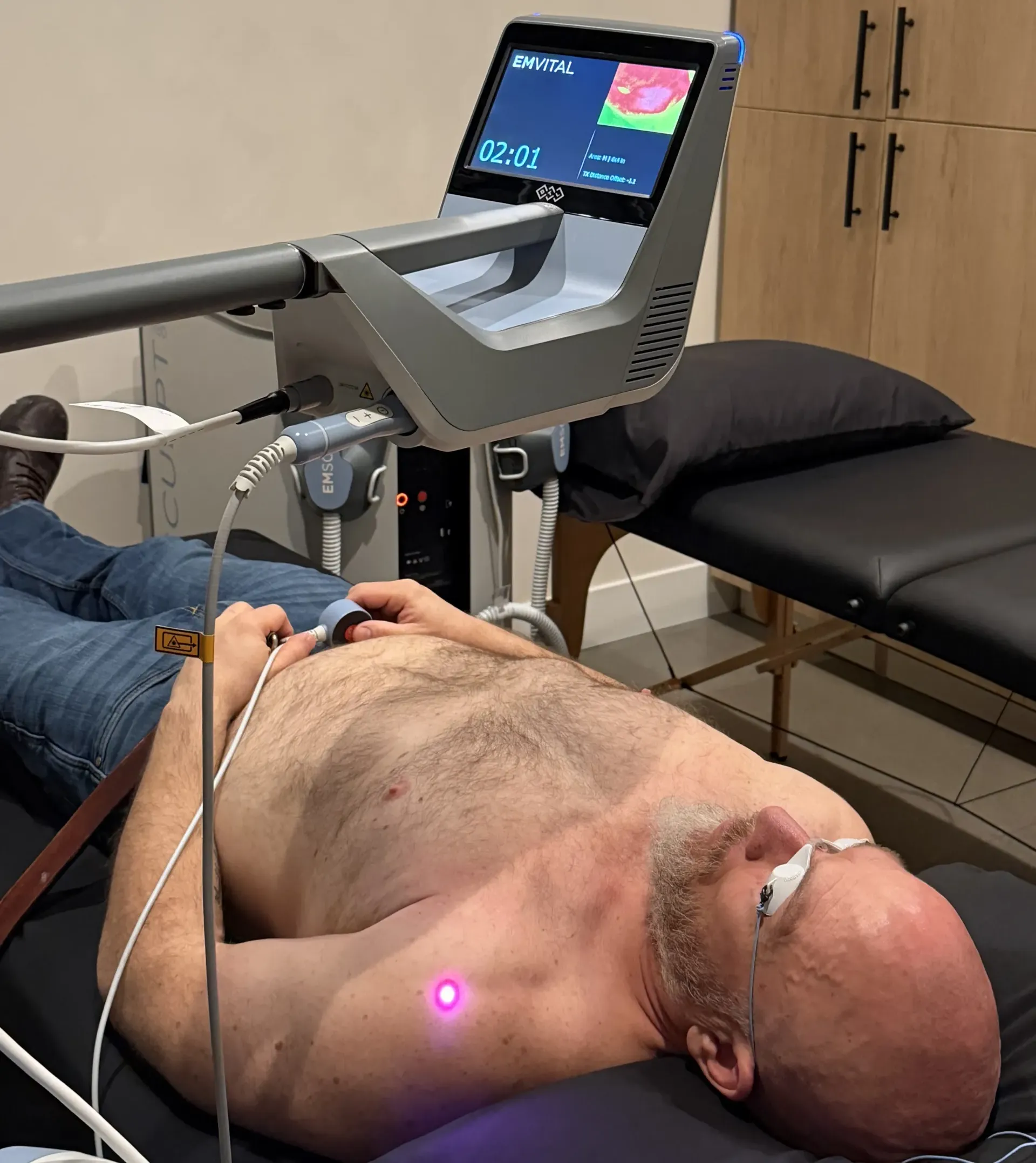 Patient lying on a medical table during a treatment using an EMVITAL device, with a purple laser on the chest area