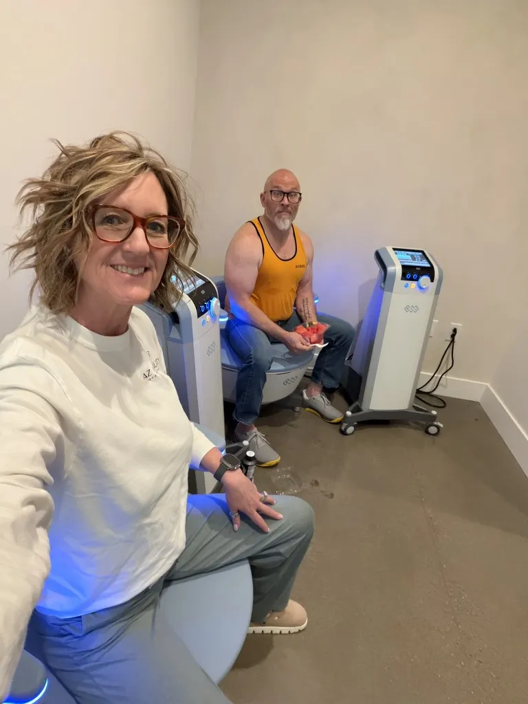 Woman with blonde hair and glasses takes a selfie in a small clinic, two futuristic blue-lit therapy chairs in the background, man in an orange tank top sits behind her, smiling at the camera.